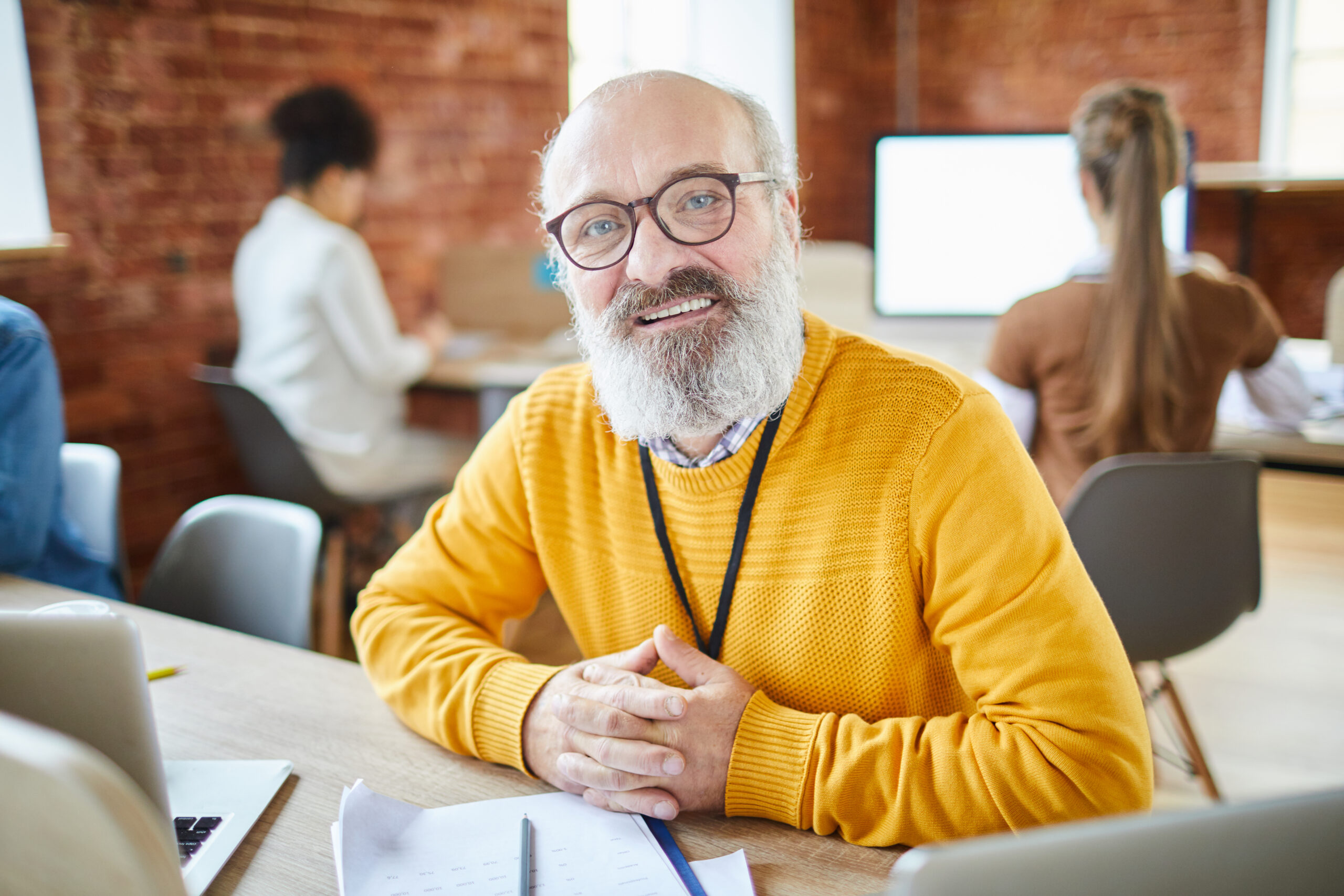 Mature confident head of business company sitting by desk with his working subordinates on background